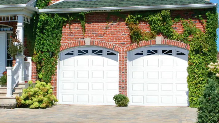 Elegant white carriage-style garage doors installed on a brick home with ivy growing around the doors, showcasing architectural integration with landscaping