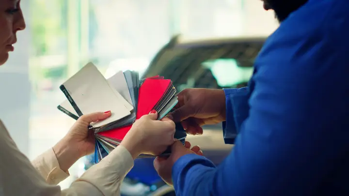 Two people hold and examine a fan of color swatches for a garage door, including a bright red sample, in front of a blurred car.
