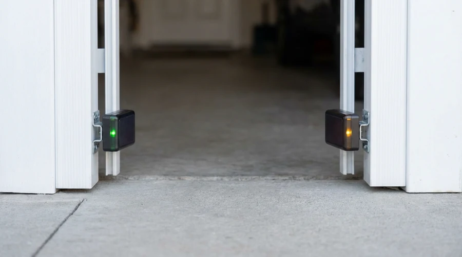 Close-up of two garage door safety sensors attached near the floor on each side of the door frame; one sensor shows a green light, the other shows an amber light.